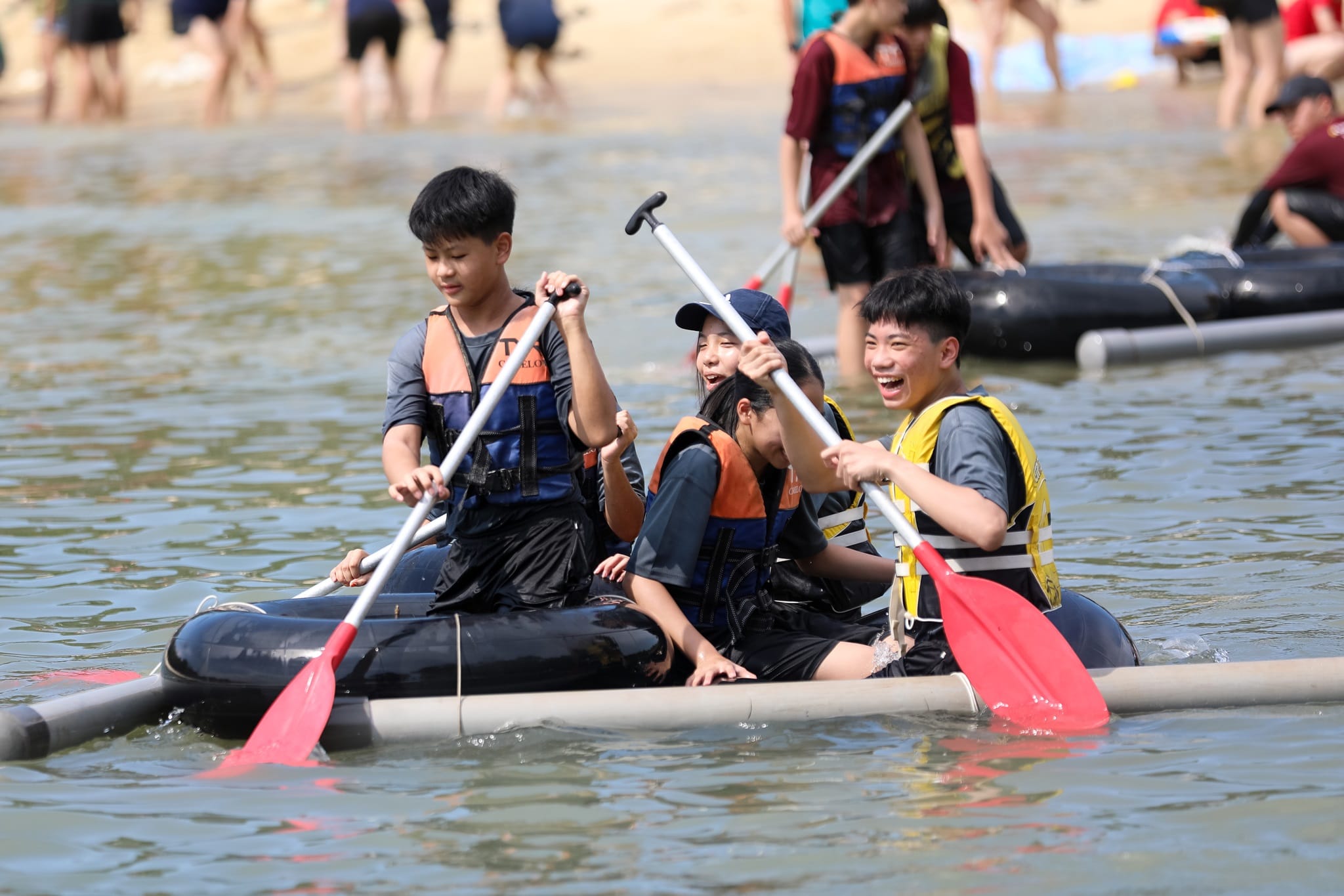 Students in life jackets smile and paddle a handmade raft together near a beach.